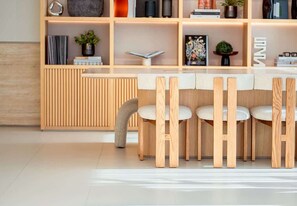 A modern interior with a wooden dining table and chairs, a bookshelf with various items, and a radiator. - Hamilton House Studio: Rooftop Pool & Gym Access (Dubai)