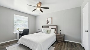 Un dormitorio con cama, escritorio, silla, ventilador de techo, ventana y puerta.