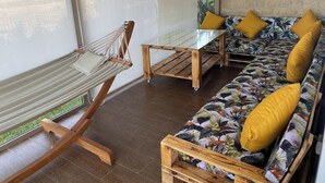 Un hamac, une table en bois et un canapé avec des coussins jaunes, le tout dans une pièce dotée de grandes fenêtres.
