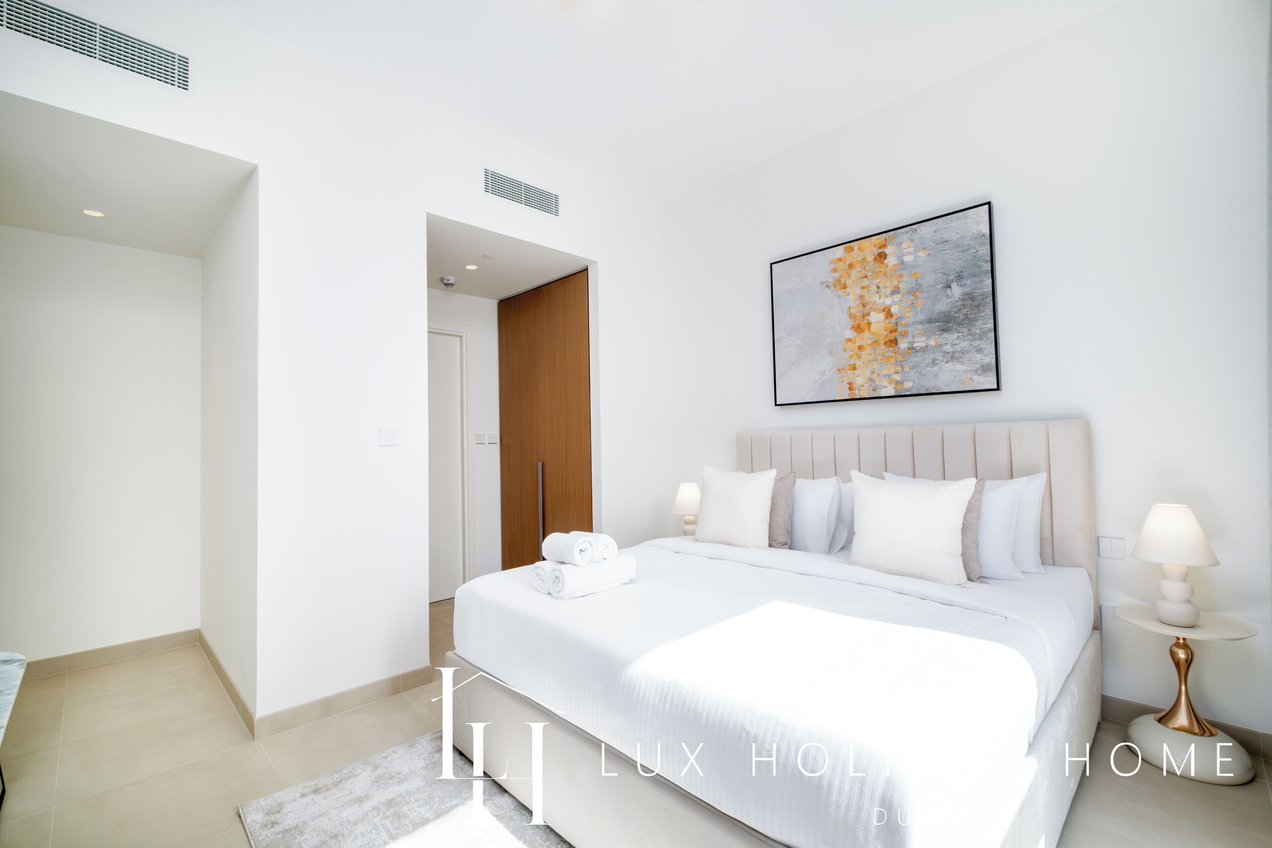 A modern hotel room with a large bed, a bedside table, a wall-mounted artwork, and a door leading to another room.