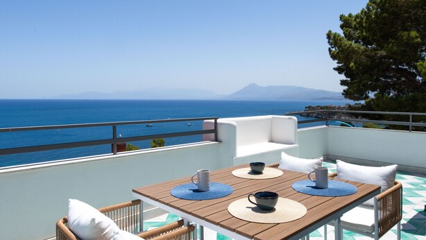 A terrace with a wooden table, white cushions, and wicker chairs overlooking a clear blue sea with boats and distant mountains.