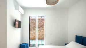 A bedroom with a bed, a TV, a blue ottoman, and a view of a stone staircase through a glass door. - Casa Solunto Vista Mare by Wonderful Italy (Santa Flavia)