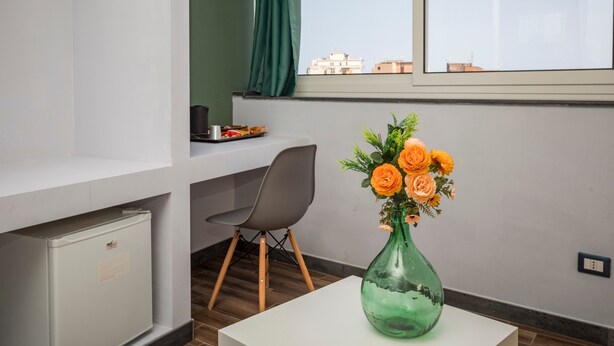 A room with a desk, a chair, a small fridge, a vase with flowers, and a window with a view of buildings. - Room 'Garibaldi - Matrimoniale' with Balcony, Wi-Fi and Air Conditioning (Catania)