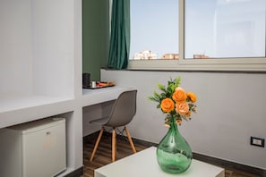 Una habitación con escritorio, silla, refrigerador pequeño, un florero con flores y ventana con vista a edificios.