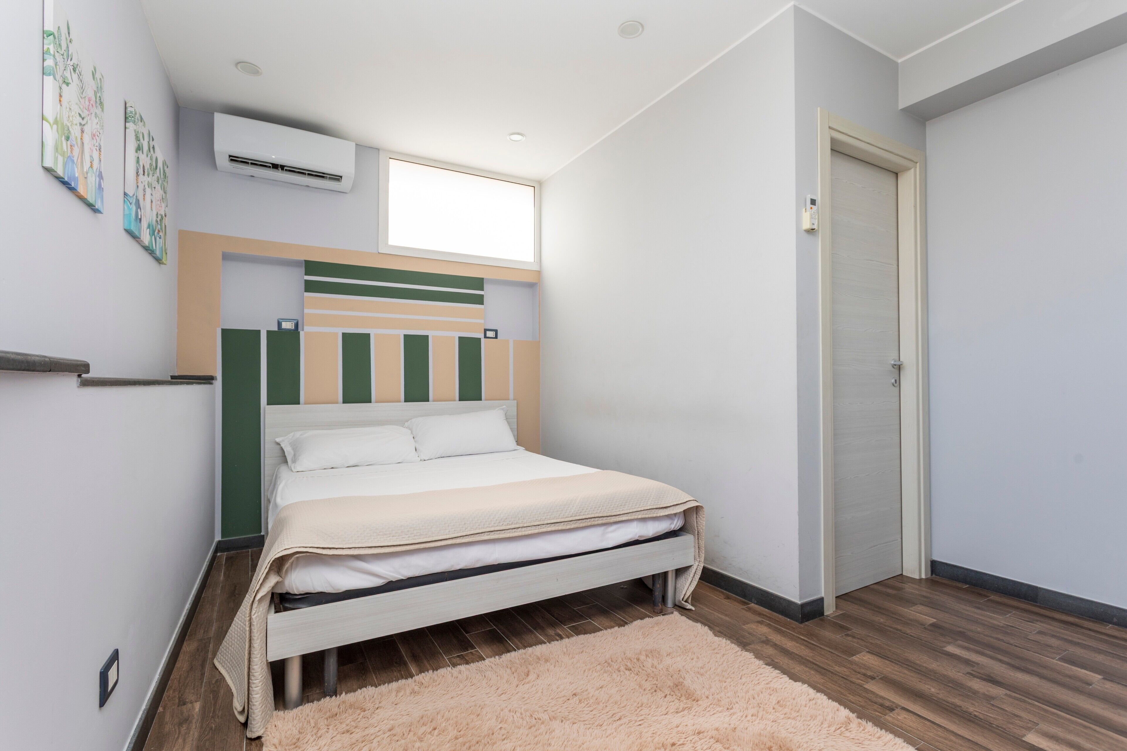 A bedroom with a bed, a window, an air conditioner, a door, and a wall-mounted shelf.