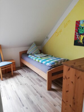Una habitación pequeña con cama individual, techo inclinado, escritorio de madera, una silla y un colorido adorno en la pared.