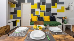 A compact room with a bed, a dining table set with plates and glasses, a small table with a vase, and a wall with geometric patterns.