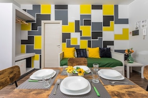 Une pièce de taille modeste comprenant un lit, une table à manger dressée avec des assiettes et des verres, une petite table sur laquelle est posé un vase, et un mur orné de motifs géométriques.