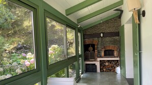 A covered outdoor area with a stone fireplace, a seating area with a white chair, and a view of a garden with pink flowers. - Flowery villa: family home, veranda and garden (Tende)