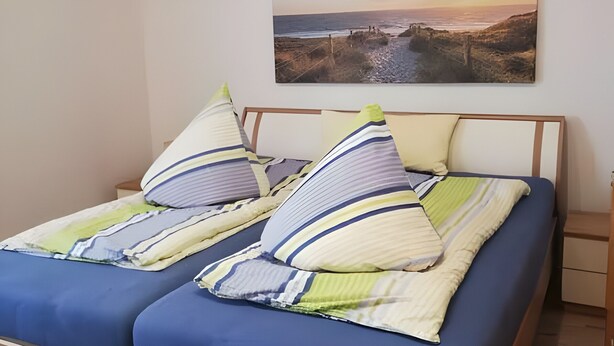 Un lit avec une literie bleue, des oreillers à rayures et un tableau représentant un paysage côtier au-dessus.