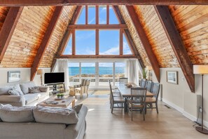A spacious loft with a high ceiling, large windows, a cozy seating area, and a dining table. - Hydrangeas by the Sea (Hyannis)