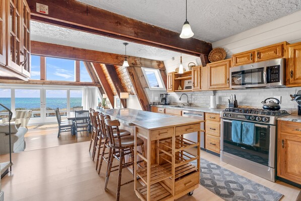 A kitchen with wooden cabinets, a large island with stools, a stove, and a view through the windows. - Hydrangeas by the Sea (Hyannis)