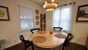 A dining area with a round wooden table, four chairs, woven placemats, a woven pendant light, a bookshelf, and a window with blinds.