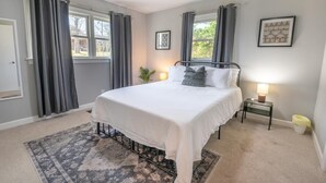 Un dormitorio con cama, ventilador de techo, mesita de noche con lámpara y ventana con cortinas.