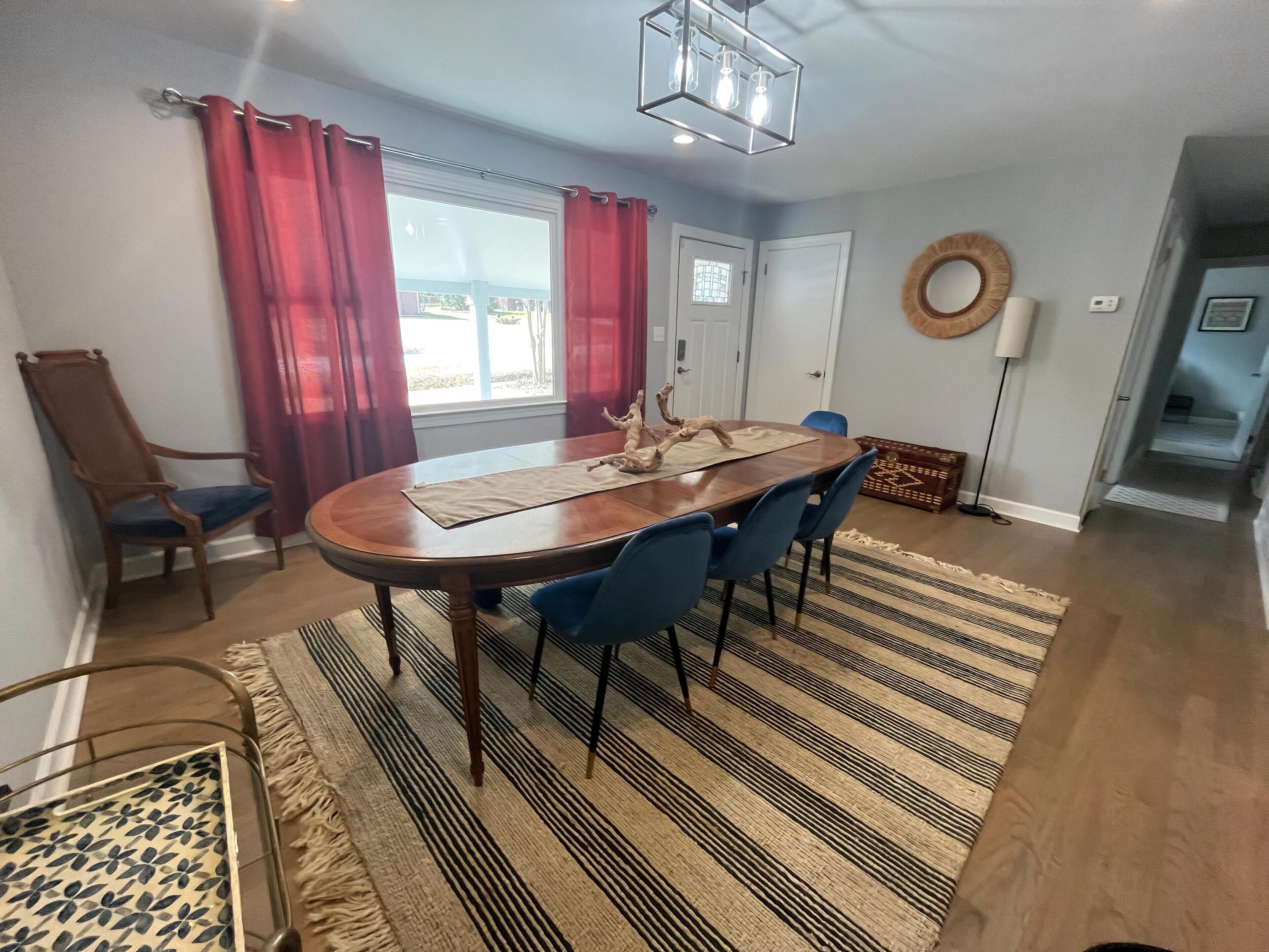 Un comedor con una mesa de madera, sillas azules, un tapete estampado y un ventanal con cortinas rojas.