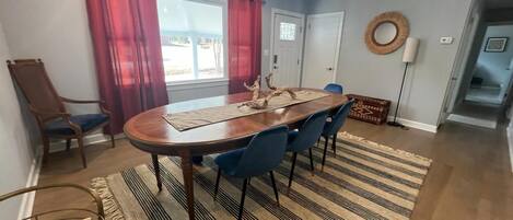 Un comedor con una mesa de madera, sillas azules, un tapete estampado y un ventanal con cortinas rojas.
