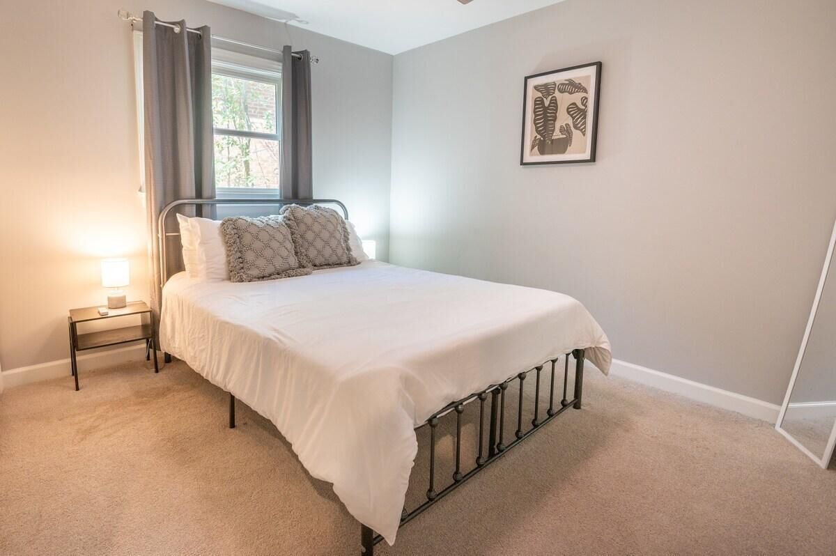 A bedroom with a bed, a bedside table, a window with curtains, and a framed picture on the wall.