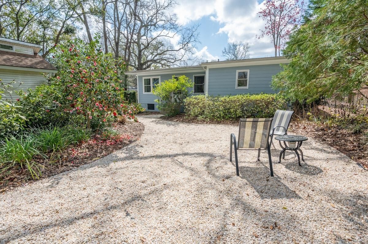 A house with a gravel driveway, a patio with a striped chair and table, and various shrubs and trees around.