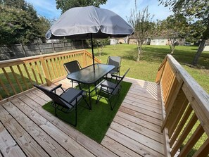 Une terrasse en bois avec une table à plateau de verre et quatre chaises, un parasol et une petite portion de gazon synthétique.