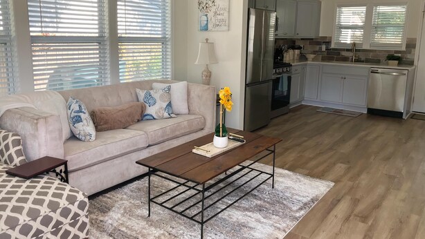 Ein Wohnzimmer mit einem beigen Sofa, gemusterten Kissen, einem Couchtisch und einem Teppich. Im Hintergrund ist eine Küche mit Edelstahlgeräten und hellen Schränken erkennbar.