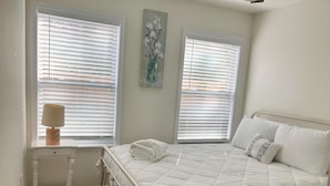 Ein Schlafzimmer mit einem Bett, zwei Fenstern mit Jalousien, einem Nachttisch mit Lampe und einer Wanddekoration.