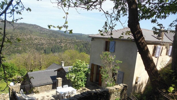 A stone wall with a stone bench and a table, a small stone building with a tiled roof, and a larger building with a tiled roof and a window with a blue shutter. - Gîte Nelly en Cévennes (Molezon)