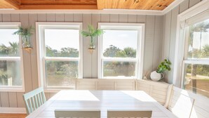 Un espace repas avec une table et des chaises en bois, de grandes fenêtres ornées de plantes et une vue sur l’extérieur.