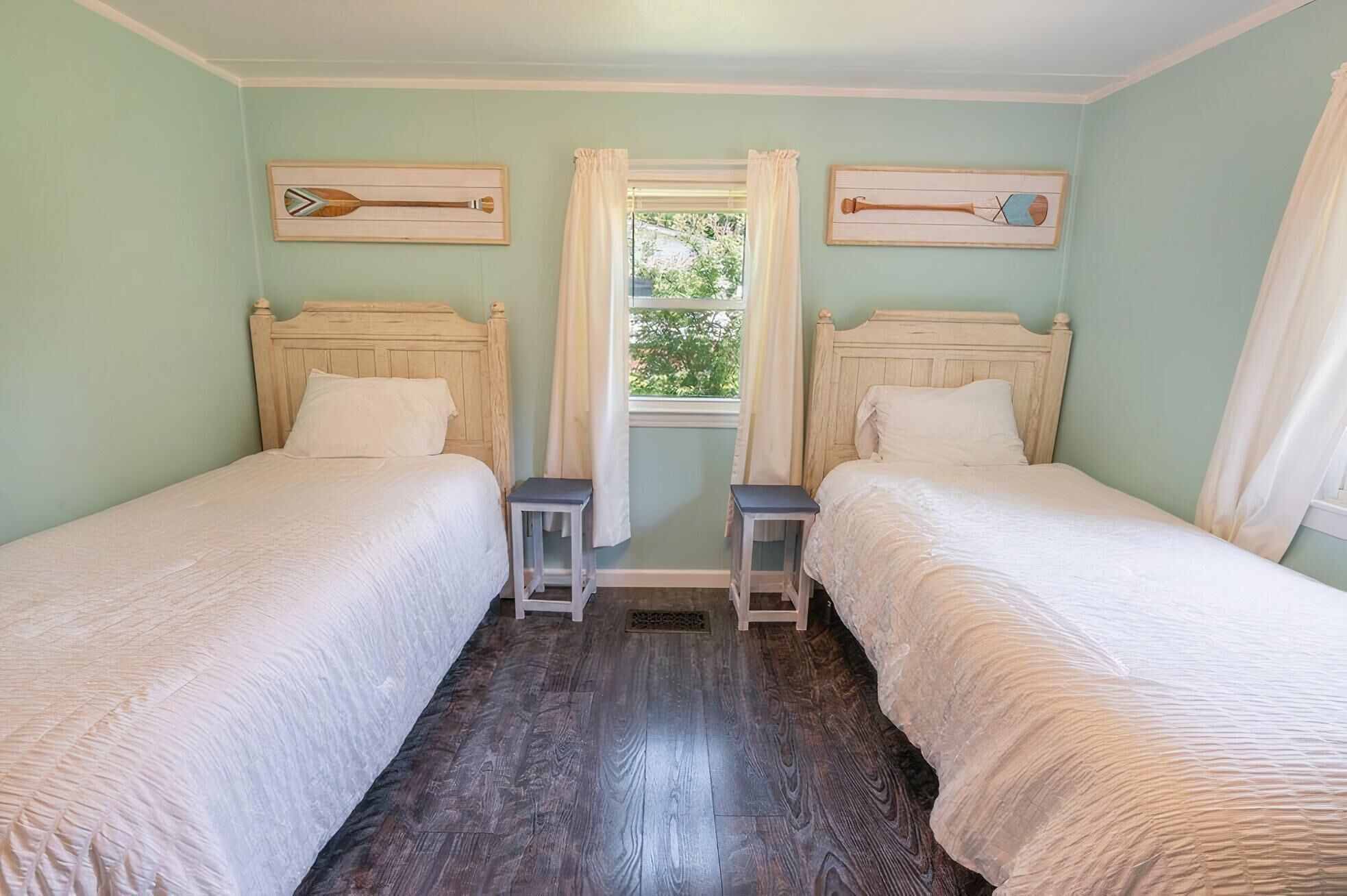 A room with two wooden beds, a window with curtains, and two wooden paddleboard paintings on the walls.