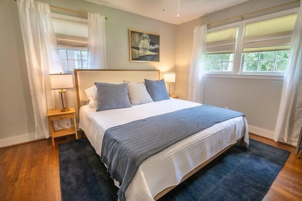 A bedroom with a large bed, two bedside tables with lamps, a painting on the wall, and a window with curtains.
