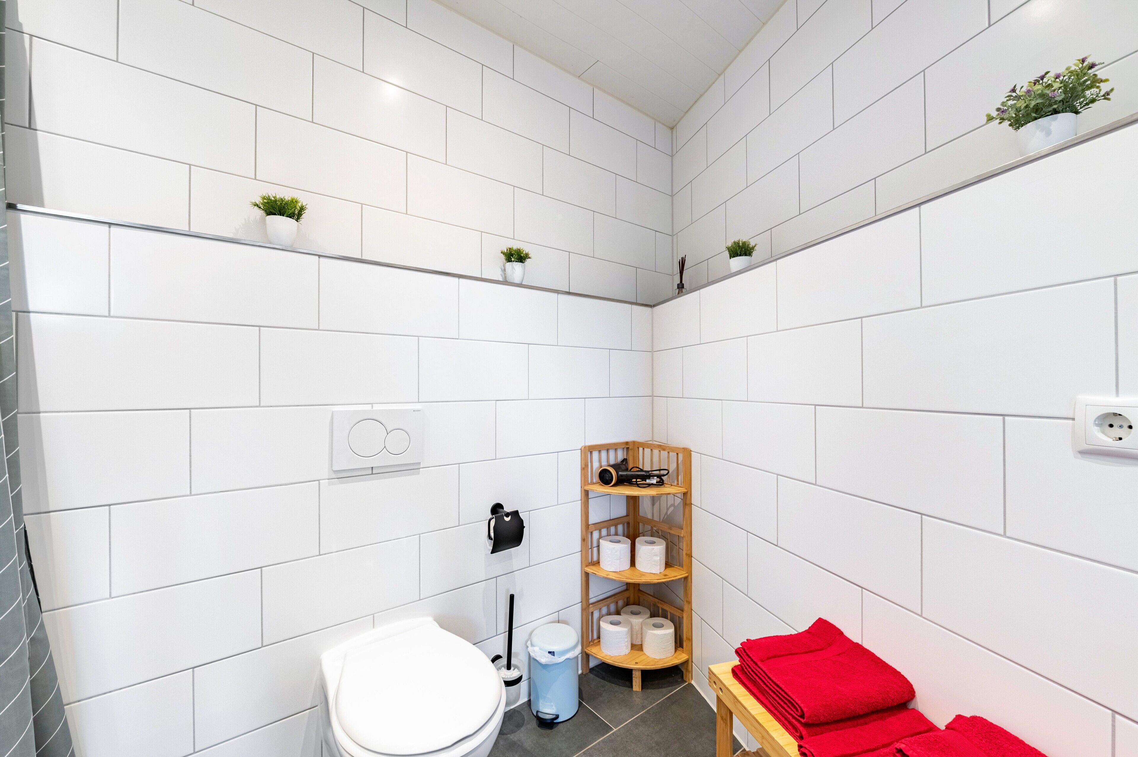 Ein kleines Badezimmer mit weißen Fliesen, einer Toilette, einem Waschbecken, einem Regal mit Pflanzen und einem Holzregal mit Handtüchern.