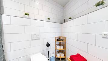 Ein kleines Badezimmer mit weißen Fliesen, einer Toilette, einem Waschbecken, einem Regal mit Pflanzen und einem Holzregal mit Handtüchern.