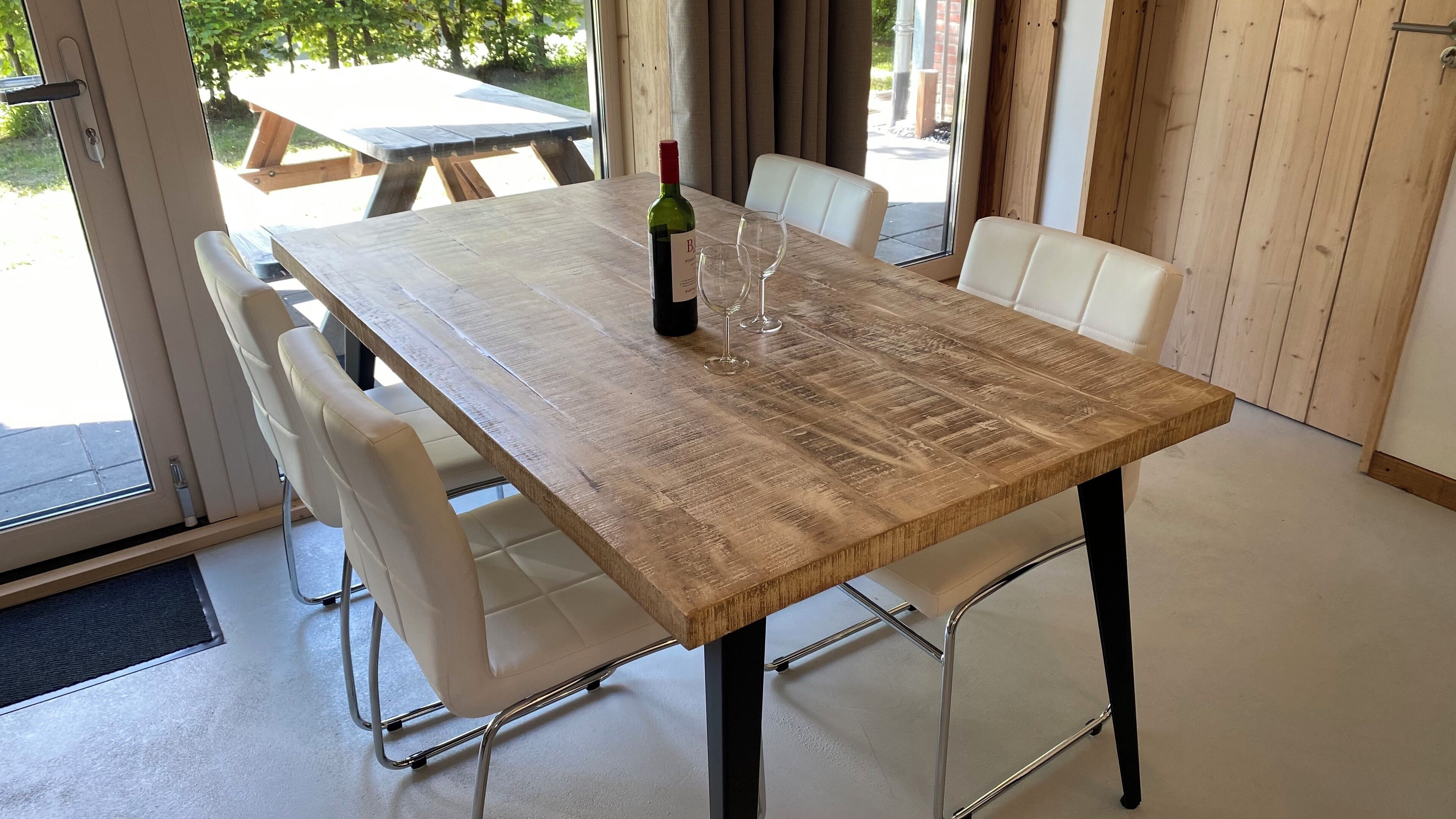 A wooden dining table with two wine glasses and a bottle, surrounded by white chairs.