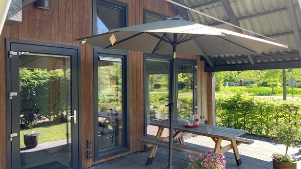 Une terrasse moderne avec une table et des chaises en bois, un grand parasol et des portes vitrées donnant sur un jardin.