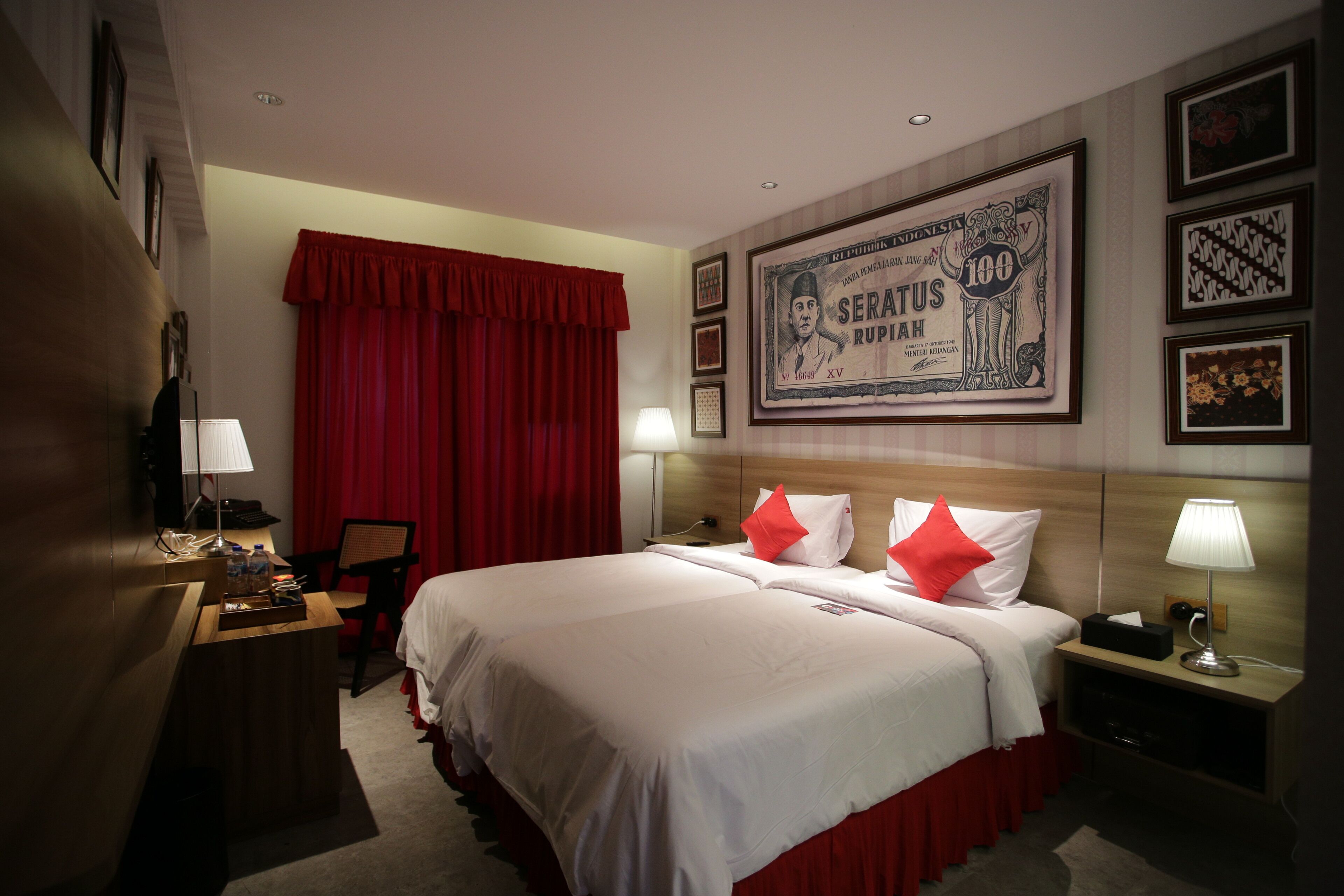 A hotel room with a large bed, red curtains, a desk with a lamp, and framed pictures on the wall.