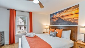 Un dormitorio ordenado con una cama grande, cabecera con una pintura de atardecer, una mesita de noche de madera y una ventana con persianas.