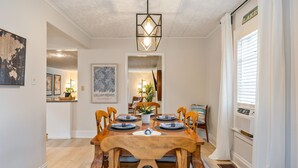 Comedor con mesa de madera dispuesta para seis personas, rodeada de sillas, lámpara colgante y cuadros en las paredes.
