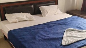 Una cama bien tendida con sábanas blancas y una manta azul, un cabecero de madera y una pared lisa con un interruptor de luz.