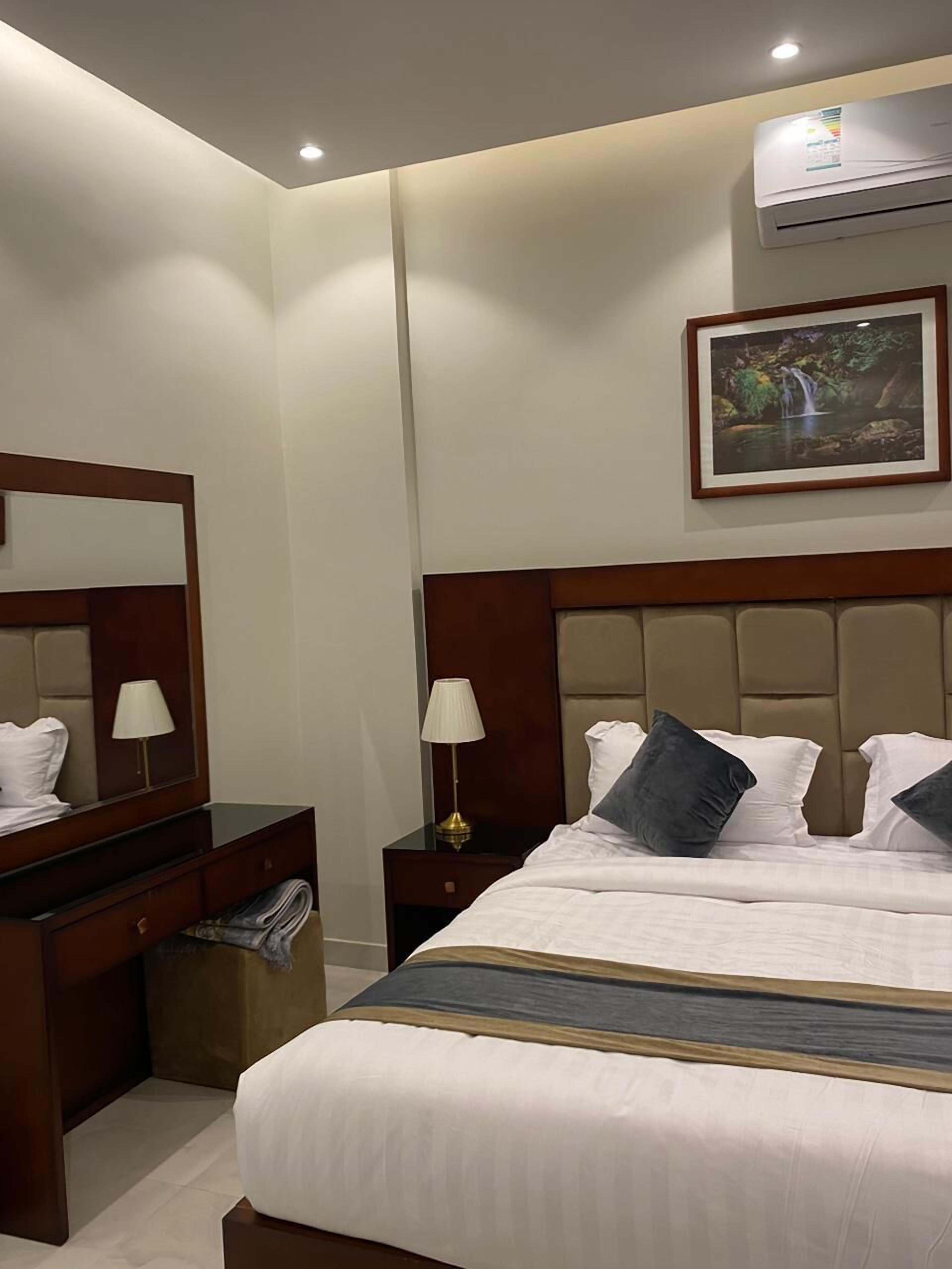 A hotel room with a large bed, a nightstand with a lamp, a dresser, a mirror, and a framed picture on the wall.