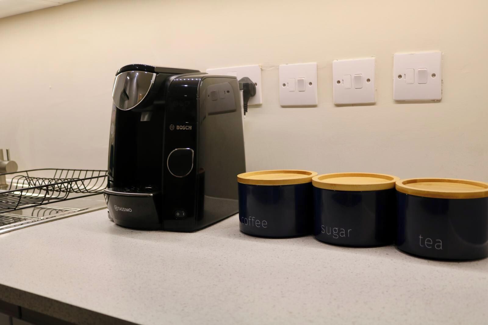 A kitchen with a coffee maker, three containers labeled coffee, sugar, and tea, and a power outlet.