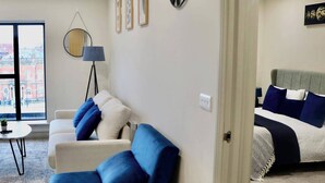 A modern living room with a sofa, a small table, and a floor lamp. There is a bed with a headboard and pillows in the adjacent room. - Cosy Urban Retreat in Leeds (Leeds)