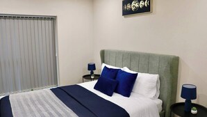 A neatly made bed with a grey upholstered headboard, white bedding, and blue pillows. There are two bedside tables with lamps, one on each side of the bed. A framed artwork depicting the moon phases hangs on the wall above the bed. - Cosy Urban Retreat in Leeds (Leeds)