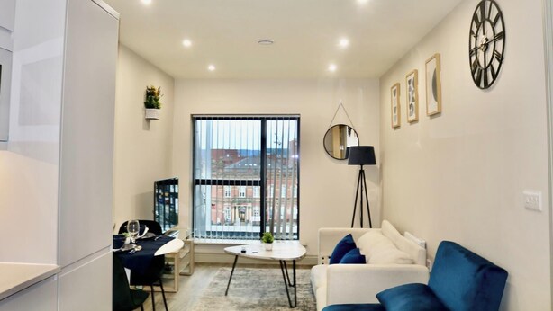 A modern living room with a white sofa, a blue armchair, a dining table with chairs, a small round table, and a large window with blinds. - Cosy Urban Retreat in Leeds (Leeds)