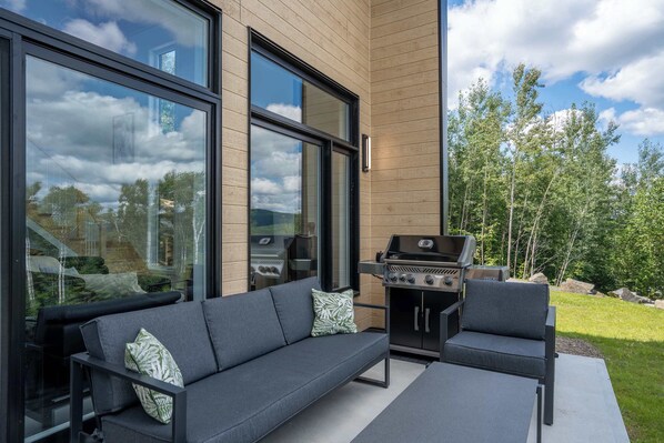 Een moderne buitenruimte met een bank en stoelen, een barbecue en grote ramen met uitzicht op het omliggende landschap.