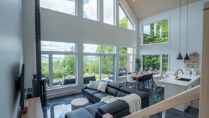 A spacious living area with a black leather sofa, a fireplace, and large windows offering a view of greenery.