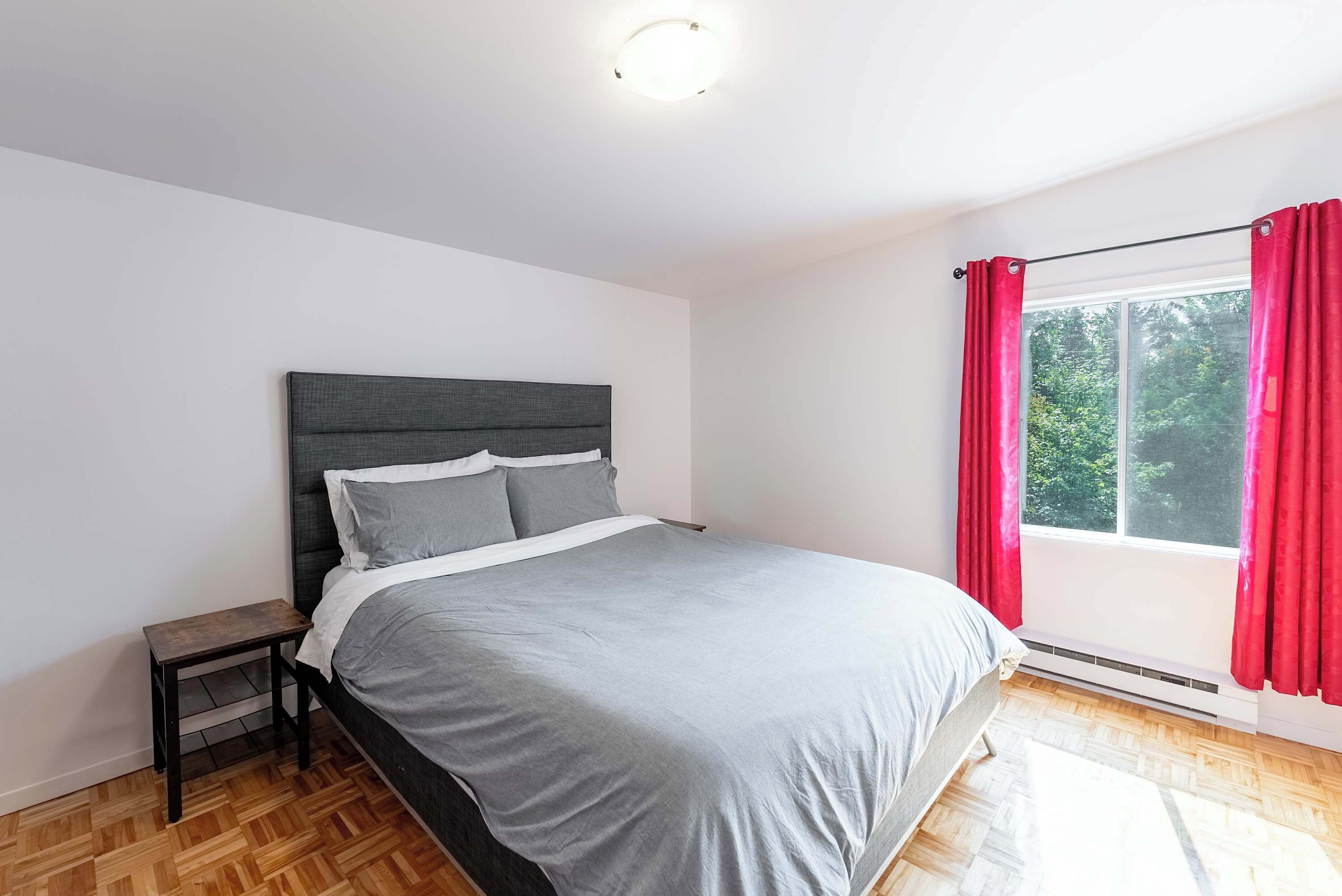 Een slaapkamer met een bed, een nachtkastje, een raam met rode gordijnen en uitzicht op groen.