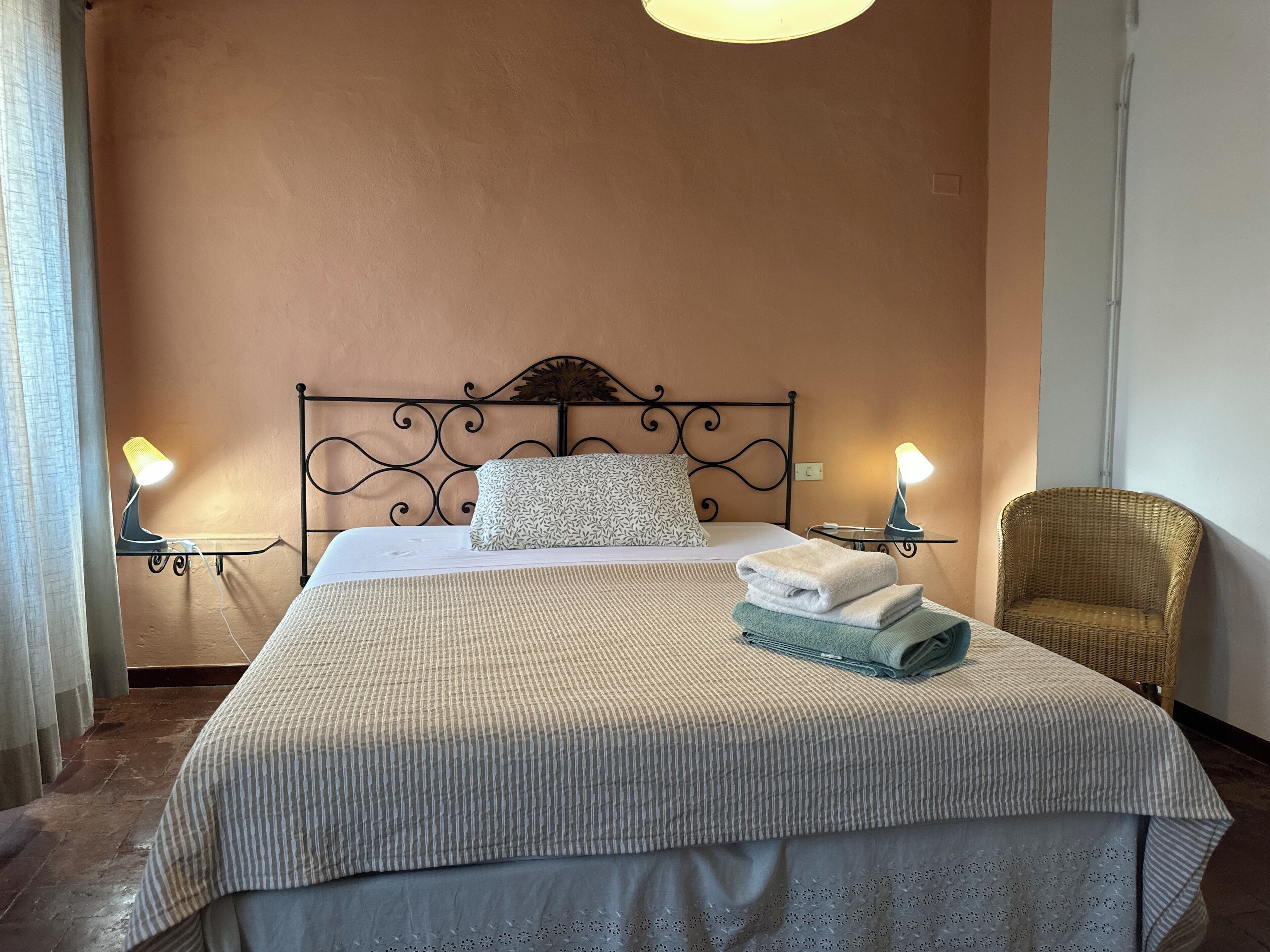 Letto ben rifatto con copripiumino a righe, due cuscini e asciugamani piegati sulla testiera. Due abat-jour su entrambi i lati del letto.