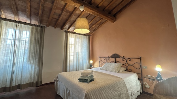 Ein Schlafzimmer mit Holzbalkendecke, einem Bett mit weißer Bettdecke, zwei Nachttischlampen und einem Korbstuhl.