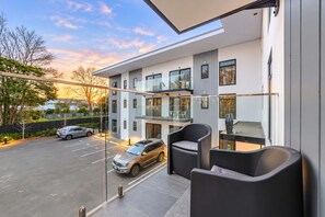 Un edificio moderno con balcón de vidrio, zona de estacionamiento y un automóvil afuera.