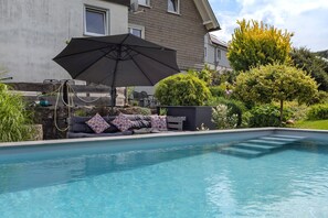 A swimming pool with a sofa and an umbrella, surrounded by greenery and a house.