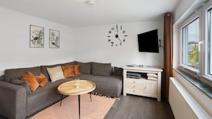 A modern living room with a grey sofa, a round wooden coffee table, a white cabinet, a mounted TV, and a large window. - Apartment 'Selbecke' with Pool and Garden (Kirchhundem)